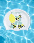 A white swim cap featuring a cute 'BEE HAPPY' design with yellow bees, a yellow honeycomb, and green leaves, shown floating on a bright blue water background with ripples.
