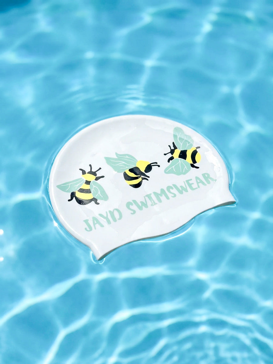 A white swim cap with printed bumblebee illustrations and the text 'JAYD SWIMSWEAR' floating on clear blue pool water.