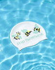 A white swim cap with printed bumblebee illustrations and the text 'JAYD SWIMSWEAR' floating on clear blue pool water.