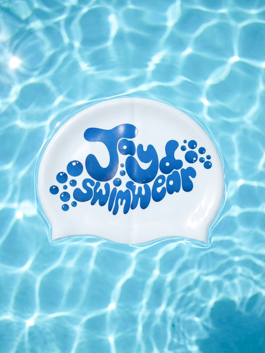 A white swim cap with blue bubble graphics and the text 'Jayd Swimwear' printed on it, floating on clear blue pool water with sunlight ripples.