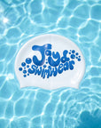 A white swim cap with blue bubble graphics and the text 'Jayd Swimwear' printed on it, floating on clear blue pool water with sunlight ripples.