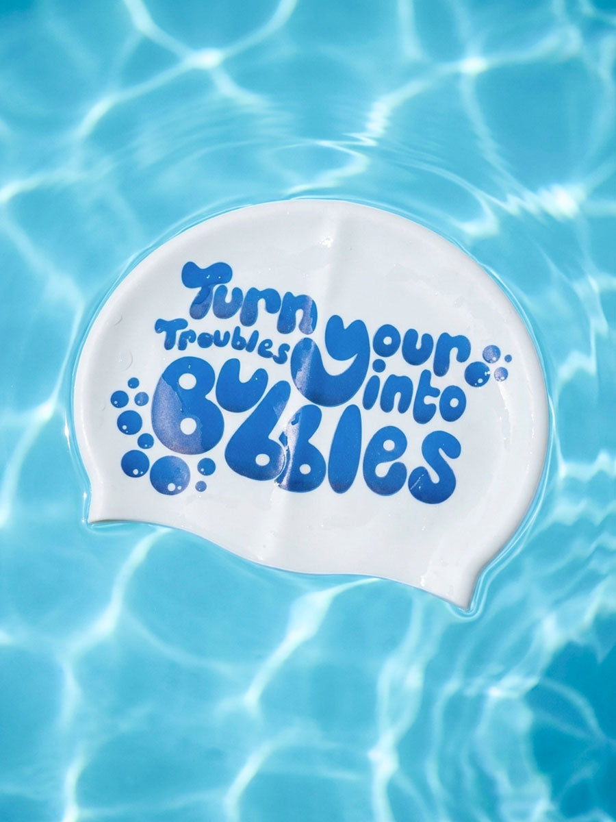 A white swim cap floating on rippling blue pool water, printed with the phrase 'Turn your troubles into bubbles' in blue bubble lettering.