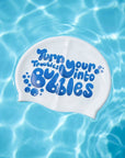 A white swim cap floating on rippling blue pool water, printed with the phrase 'Turn your troubles into bubbles' in blue bubble lettering.