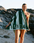 JAYD Swimwear model Emily standing on a sandy beach in front of rocky cliffs, wrapped in a green and white striped beach towel with the name 'JAYD' and a small JAYD Gem printed on it.