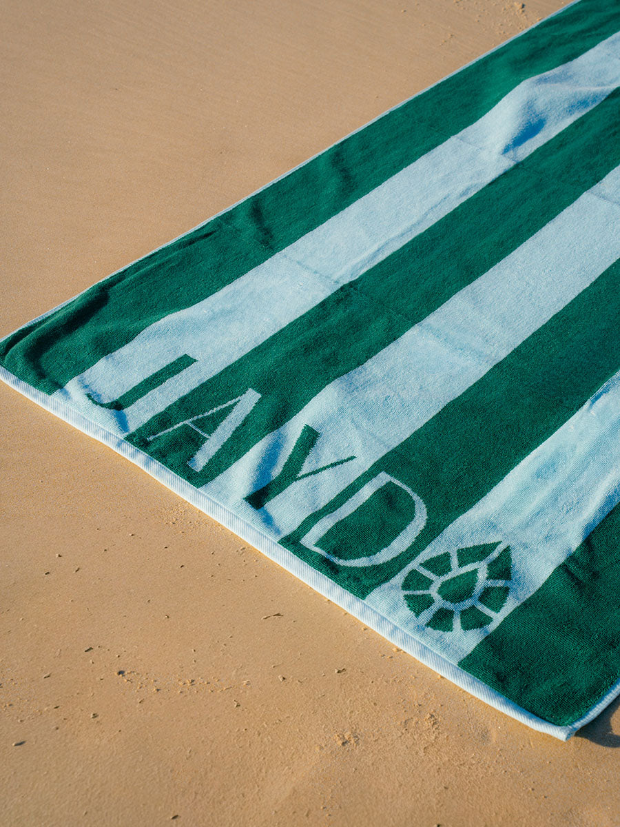 A green and blue striped beach towel laid diagonally on sandy ground, with the  'JAYD' printed in large letters along the bottom with the JAYD Gem icon near the corner.