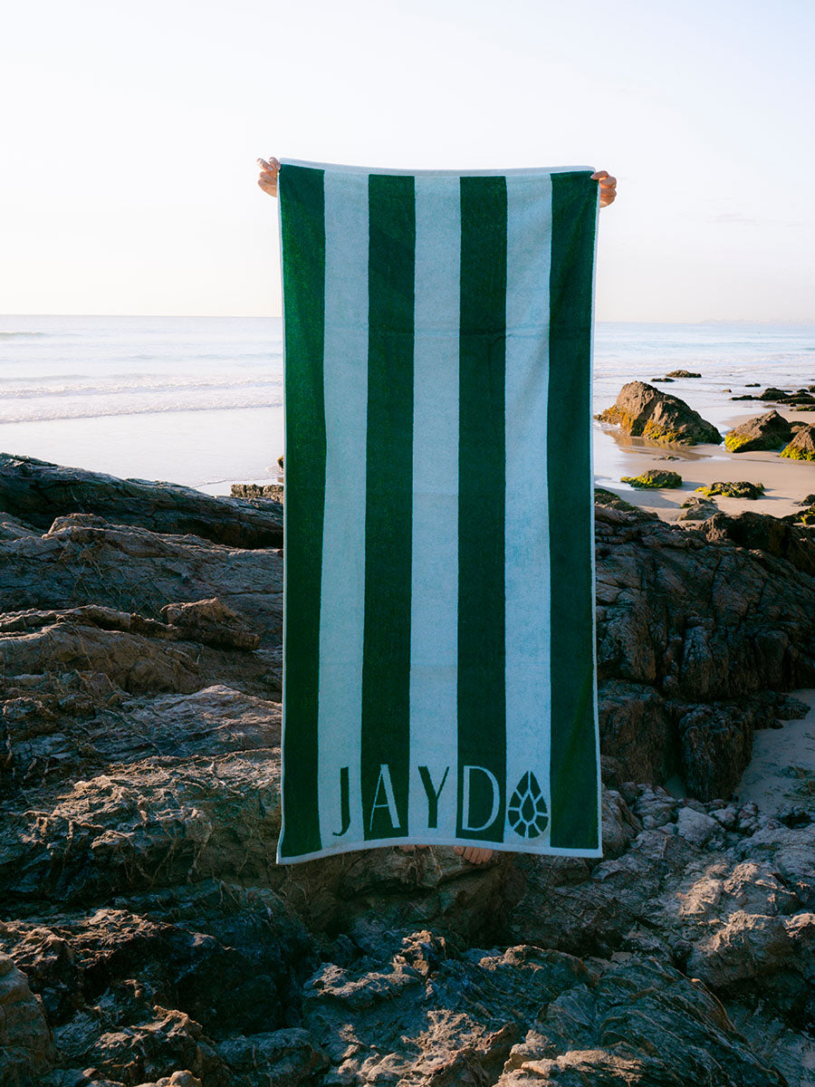 A person holds up a large beach towel with vertical green and white stripes on a rocky beach with the ocean in the background. The towel has the name 'JAYD' and a small JAYD Gem logo printed near the bottom.