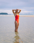 JAYD Swimwear model Kate wearing the Blaze Lennox one-piece swimsuit with contrasting cacao shoulder straps and hip cut-outs standing in shallow water at a beach, with her hands on her head, and a distant city skyline and island visible in the background.