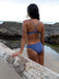 Athlete Ana standing on a beach leaning on a log staring out at the ocean, wearing the JAYD Swimwear Cali bottoms in Storm blue.