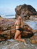 Model Chloe standing in front of a rock at the beach, wearing the mid-coverage JAYD Cali bottoms in black.