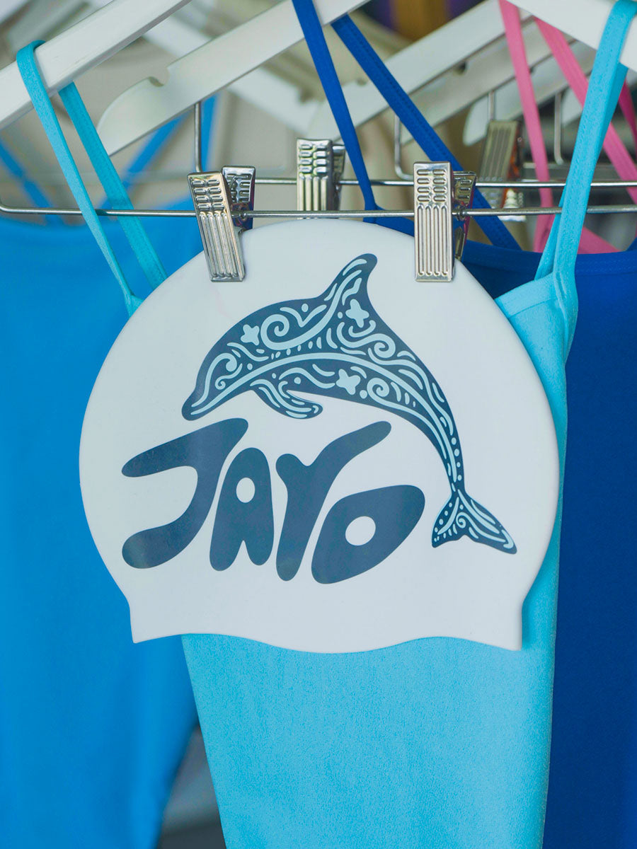 White swim cap with dolphin design and 'JAYD' branding hanging on a rack of complimentary blue JAYD one piece swimsuits.