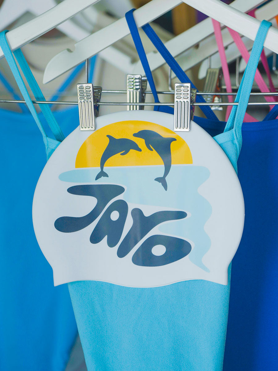 White swim cap with dolphin jumping out of the water at sunrise and 'JAYD' text branding, hanging on a rack of complimentary blue JAYD one piece swimsuits.