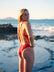 JAYD athlete Mia Cook standing on a rocky beach looking over her shoulder wearing the Finley one piece swimsuit in Ruby red.