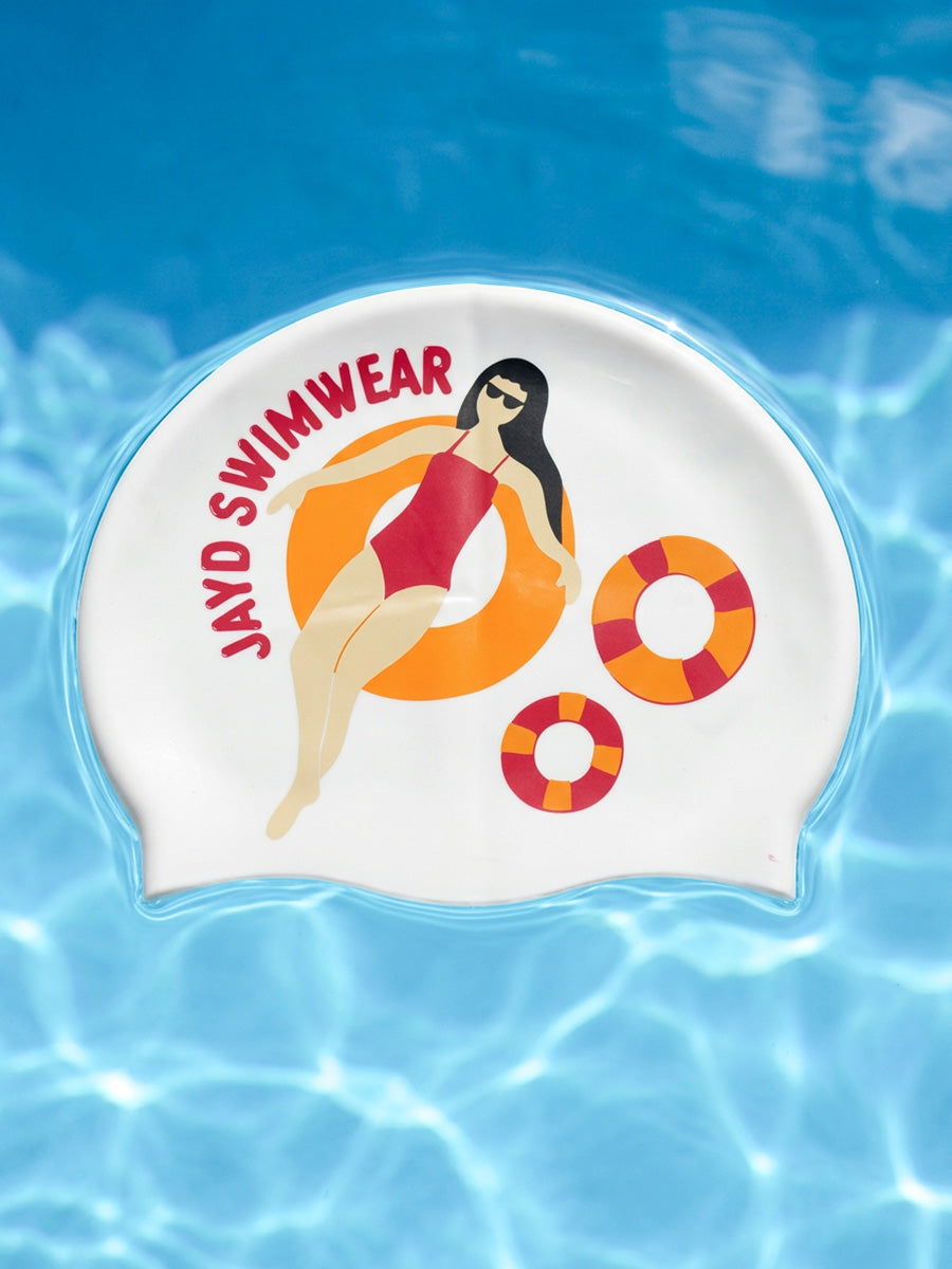 A white swim cap featuring a cartoon illustration of a woman in a red swimsuit lounging on an orange float, with the text 'JAYD SWIMWEAR' in red. The swim cap is shown floating on a bright blue water background with ripples.
