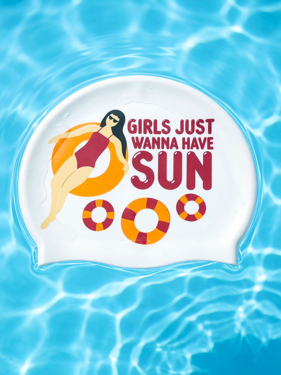 A white swim cap with a colorful graphic of a woman on an orange float and the text 'GIRLS JUST WANNA HAVE SUN' is shown floating on blue pool water.