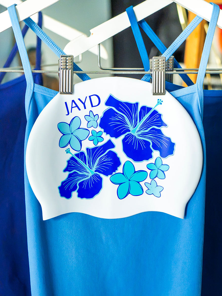 White swim cap hanging on a rack with blue hibiscus flowers, matching JAYD Swimwear's blue swimsuit range.
