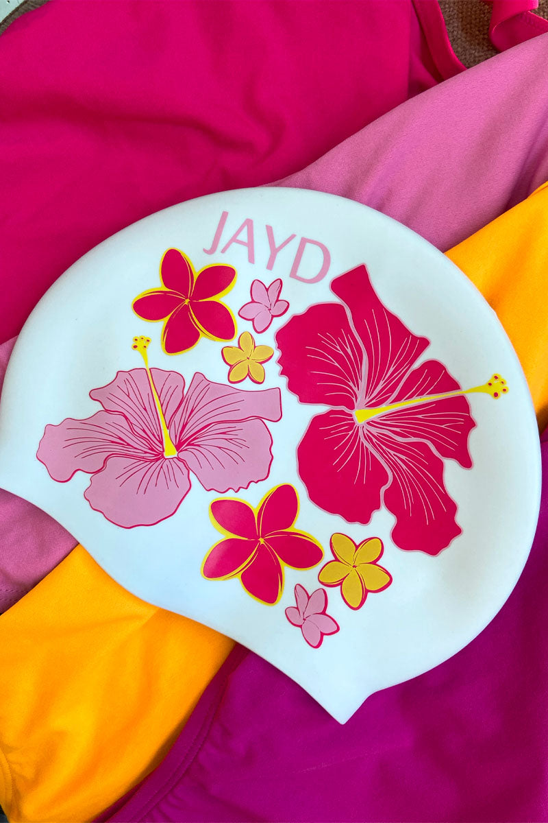 White Hibiscus Haven swim cap sitting on pink and orange JAYD one piece swimsuits, featuring pink and orange flowers.