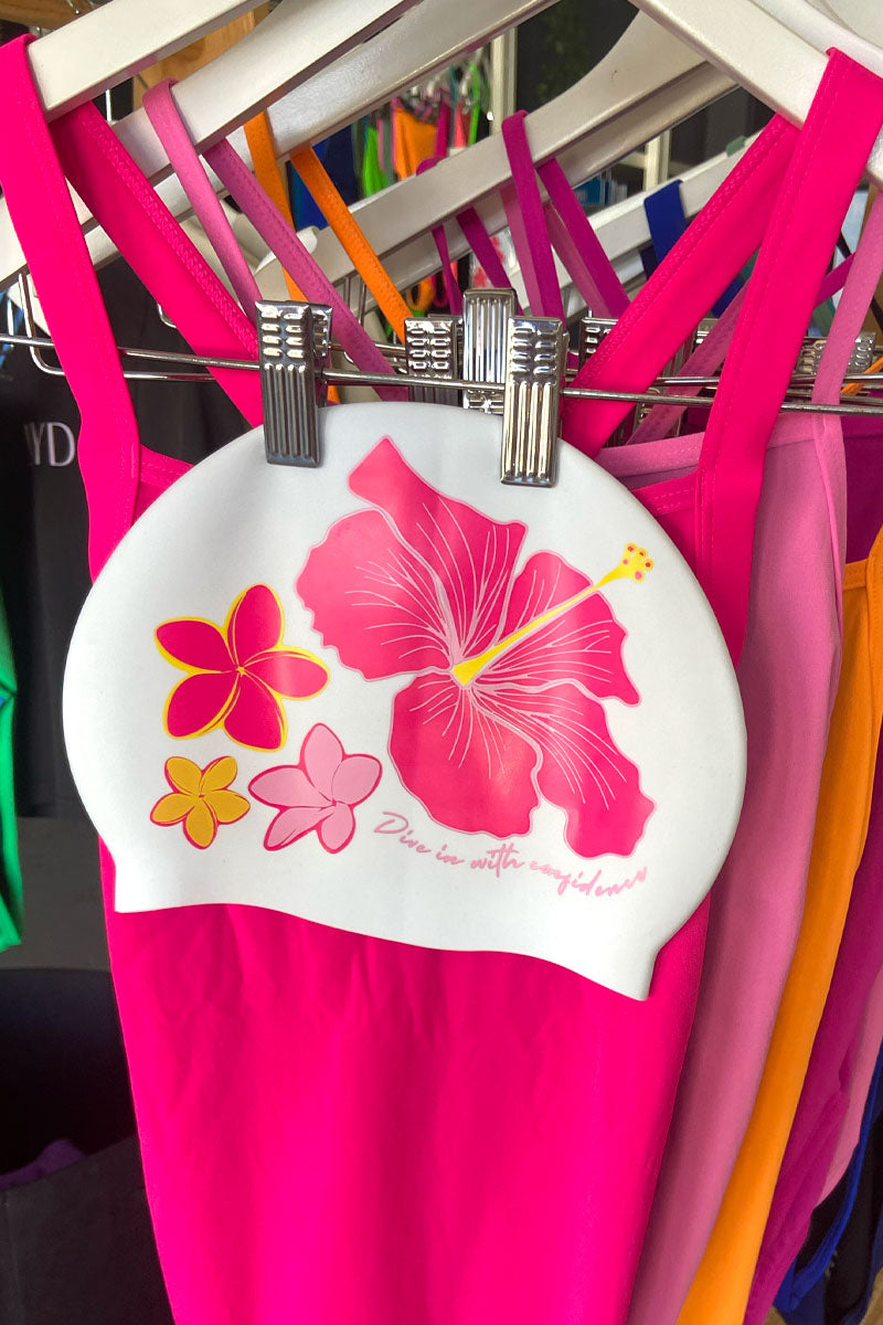 White swim cap with pink floral hibiscus design hanging on a rack with colourful pink and orange JAYD Swimwear one piece swimsuits.