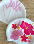 Pink JAYD swim caps Shaka and Hibiscus Haven on a stone background.