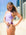 JAYD Swimwear model Emily standing on the beach with tall buildings in the distance, adjusting the chubby cut-out hip details on her Lilac Haze x Sangria Ibiza one pice swimsuit.