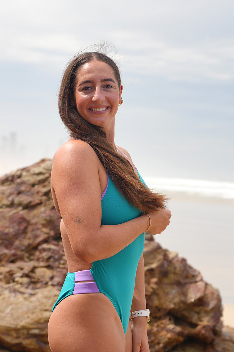 Ski Paddler Jenaya Massie on the beach wearing the JAYD Swimwear Ibiza One Piece in Atlantic with Lilac Haze straps & contrasting hip cut-outs.