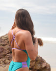 A close-up snapshot of bold lilac haze hip cut-outs contrasting against a vibrant atlantic Ibiza JAYD swimsuit body as model looks off into the distance of the beach with waves crashing agains the shoreline.