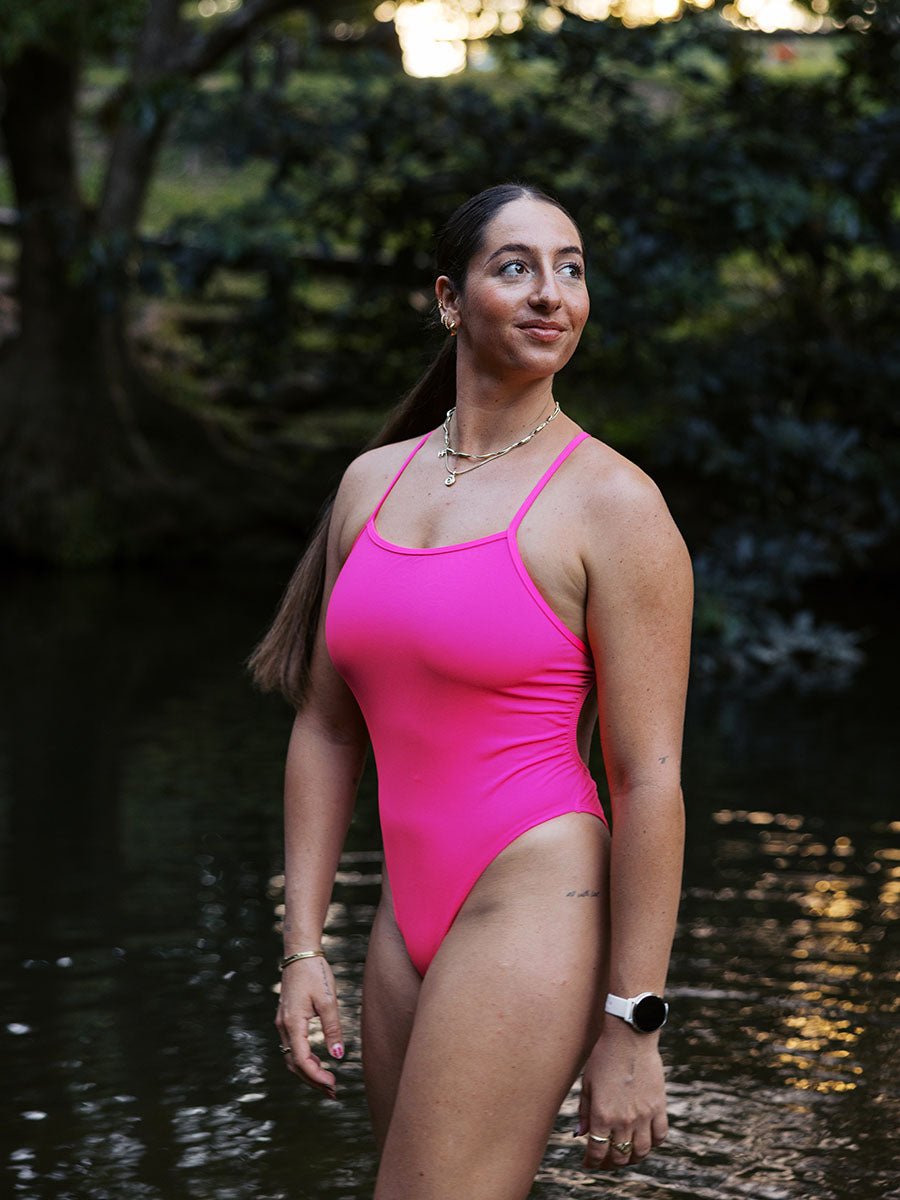 Athlete Jenaya standing in a river with a green forest behind her, wearing the JAYD Isla one piece in pink sorbet.