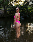 Athlete Jenaya showcasing the back and side view of the JAYD Swimwear Isla one piece tie back straps standing in water with trees and foliage in the background.