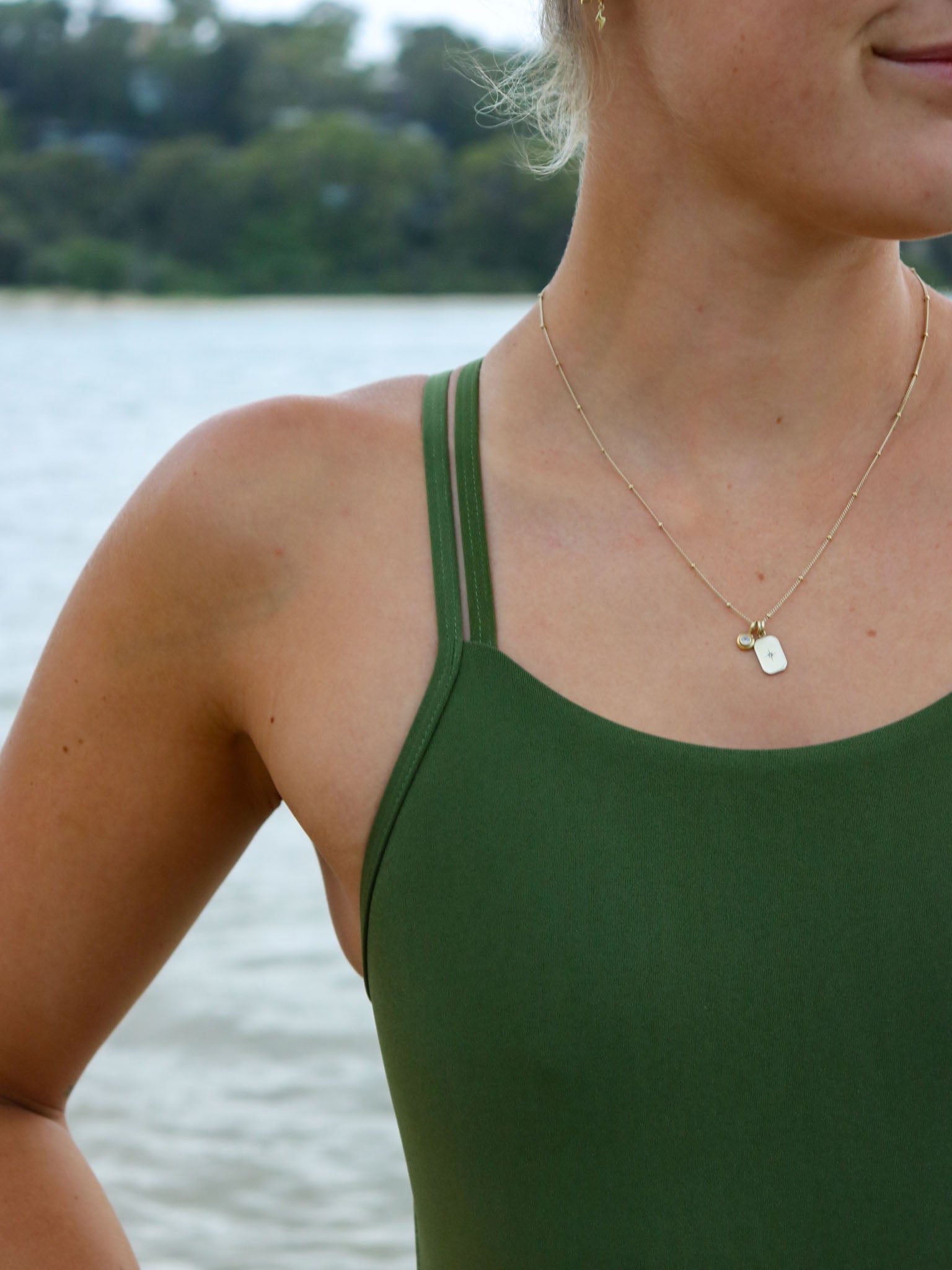Close shoulder strap view of a female athlete wearing a JAYD Swimwear athletic one piece swimsuit, Apsley in Olive green, standing on a beach.