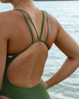 Close back strap view of a female athlete wearing a JAYD Swimwear athletic one piece swimsuit, Apsley in Olive green, standing on a beach.
