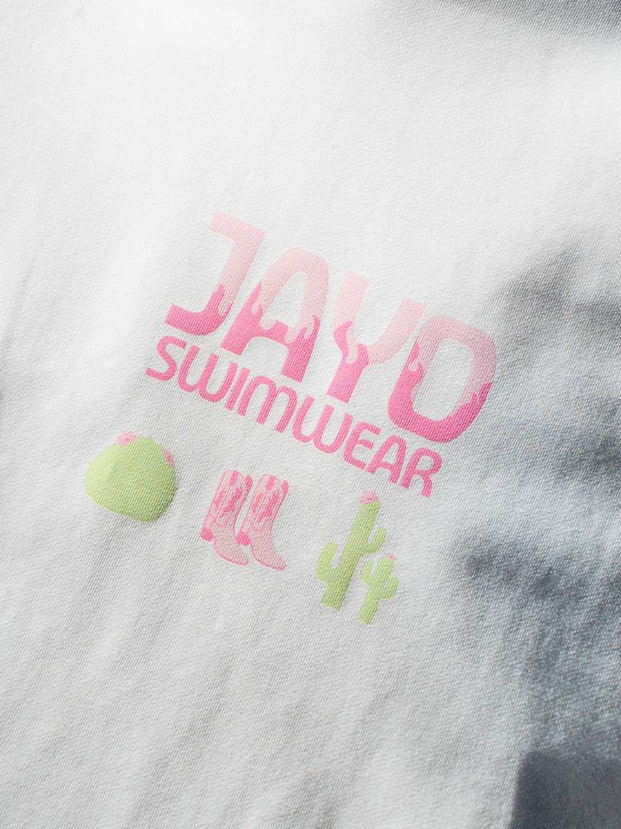 Fabric close-up of JAYD Swimwear's white oversized tee, printed with a pink JAYD Swimwear word mark and a western-inspired cactus and boots motif.