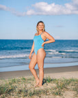 Athletic one piece by JAYD swimwear in Waikiki blue with Cancun accent straps, standing by the ocean

