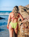 Active fixed back one piece swimsuit in Kiwi green with Tart pink accents, standing on rocky shoreline. Featured suit is the Zambia one piece by JAYD Swimwear.