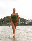 Front view of a female athlete walking on a beach while wearing a JAYD Swimwear one piece swimsuit, Lennox in Olive green with blue hip cut outs.