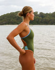 Side view of a female athlete on a beach wearing a JAYD Swimwear one piece swimsuit, Lennox in Olive green with blue hip cut outs.