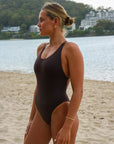 Front view of a woman in a JAYD Swimwear Levanto one piece swimsuit in cacao, standing on a sandy beach with water and trees in the background.
