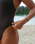 Close hip view of a woman wearing a seamless JAYD Swimwear Levanto one piece swimsuit in cacao, standing on a sandy beach with water and trees in the background.