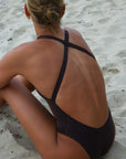 Back strap view of a woman in a JAYD Swimwear Levanto one piece swimsuit in cacao, sitting on a sandy beach.