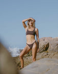 Woman in a brown JAYD Swimwear bikini top, Nalu in Cacao, standing on rocks by the ocean with a clear blue sky.
