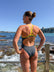 JAYD athlete, Lizzie Welborn, wearing a dark gray one-piece swimsuit with neon yellow straps, photographed from behind while standing on a rocky seaside promenade with the ocean and blue sky in the background.