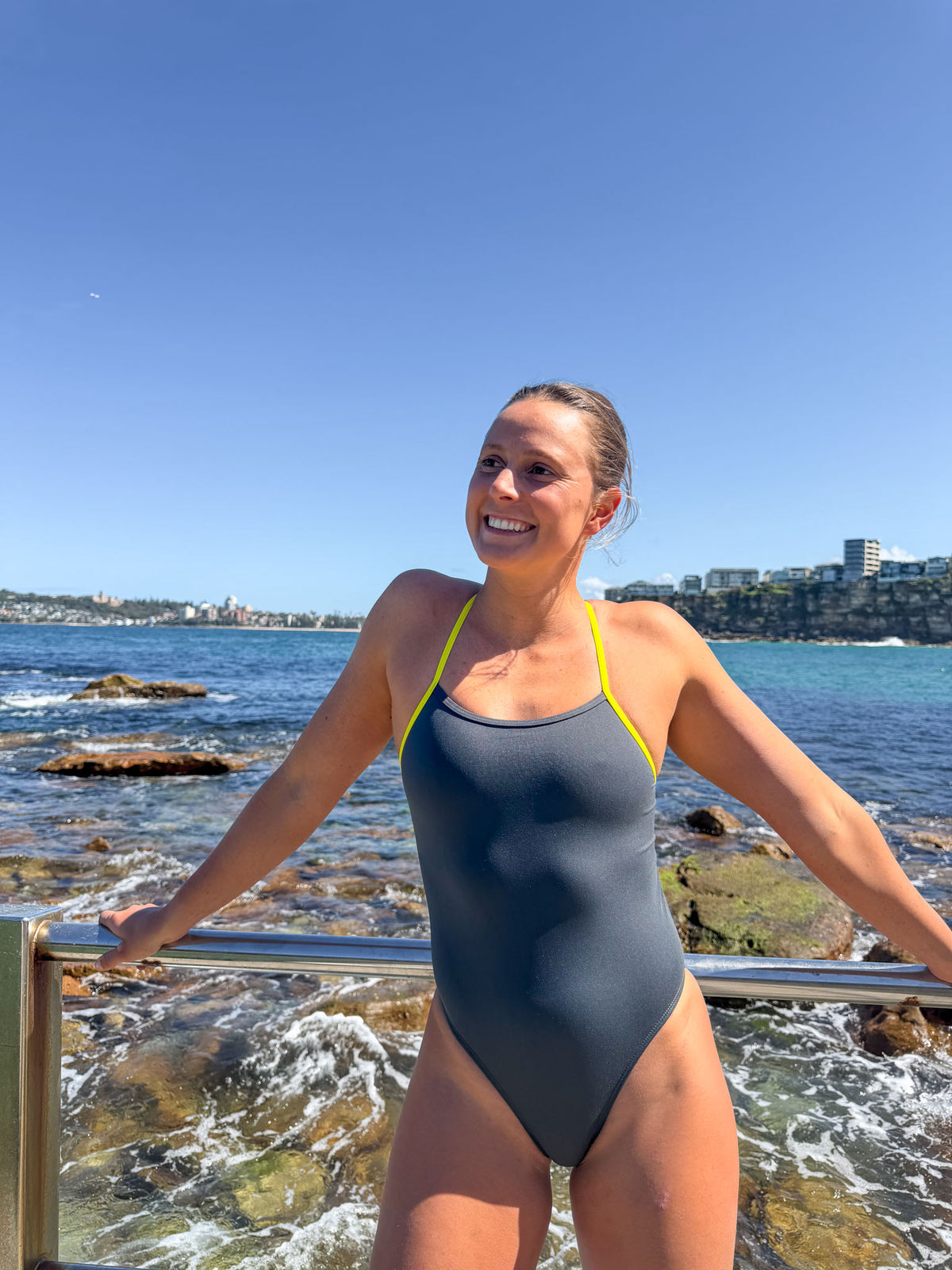 JAYD athlete, Lizzie Welborn, wearing a dark grey one-piece swimsuit with bright yellow straps stands on a rocky beach with the ocean and blue sky in the background.