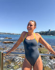 JAYD athlete, Lizzie Welborn, wearing a dark grey one-piece swimsuit with bright yellow straps stands on a rocky beach with the ocean and blue sky in the background.