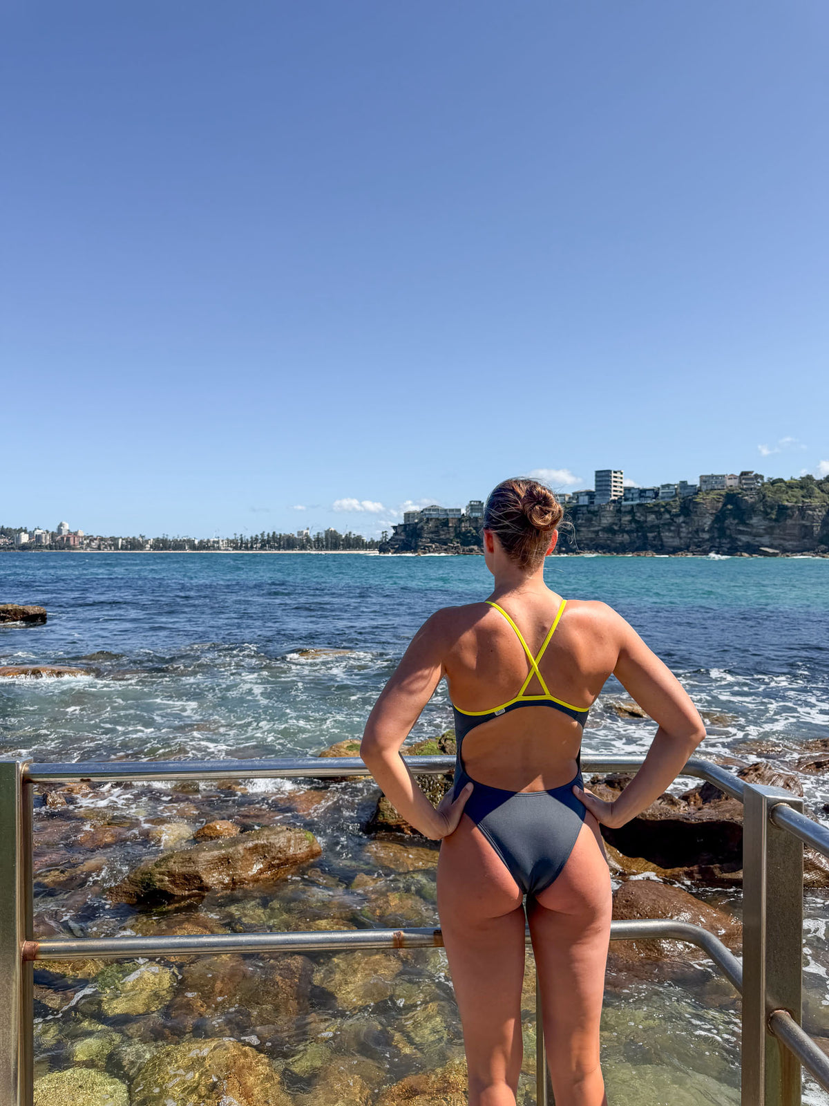 JAYD athlete, Lizzie Welborn, stands with her back to the camera on a rocky coastal walkway, wearing a dark gray carbon JAYD Swimwear siena one-piece swimsuit with a bright yellow-green crisscross back strap. She is looking out over the blue ocean and distant cliffs under a clear blue sky.