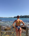 JAYD athlete, Lizzie Welborn, stands with her back to the camera on a rocky coastal walkway, wearing a dark gray carbon JAYD Swimwear siena one-piece swimsuit with a bright yellow-green crisscross back strap. She is looking out over the blue ocean and distant cliffs under a clear blue sky.