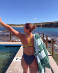JAYD athlete, Lizzie Welborn, stands on a concrete poolside deck with her back to the camera, wearing a carbon gray JAYD Swimwear Siena one-piece swimsuit with bright yellow strappy details on the back. She has a green and white JAYD towel draped over her shoulder, with a rocky coastline and blue ocean in the background.