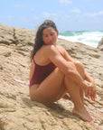 Side view of female swimmer sitting on a rock wearing JAYD Swimwear athletic one piece swimsuit, Soraya in Sangria.
