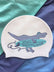 A white swim cap featuring a cartoon T-rex holding a teal surfboard with the text 'JAYD SWIMWEAR', displayed on a background of teal and navy blue fabric.