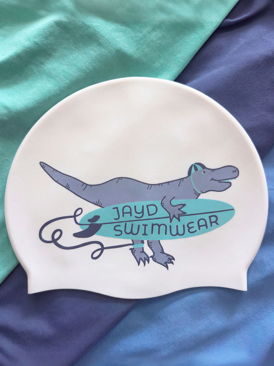 A white swim cap featuring a cartoon T-rex holding a teal surfboard with the text 'JAYD SWIMWEAR', displayed on a background of teal and navy blue fabric.