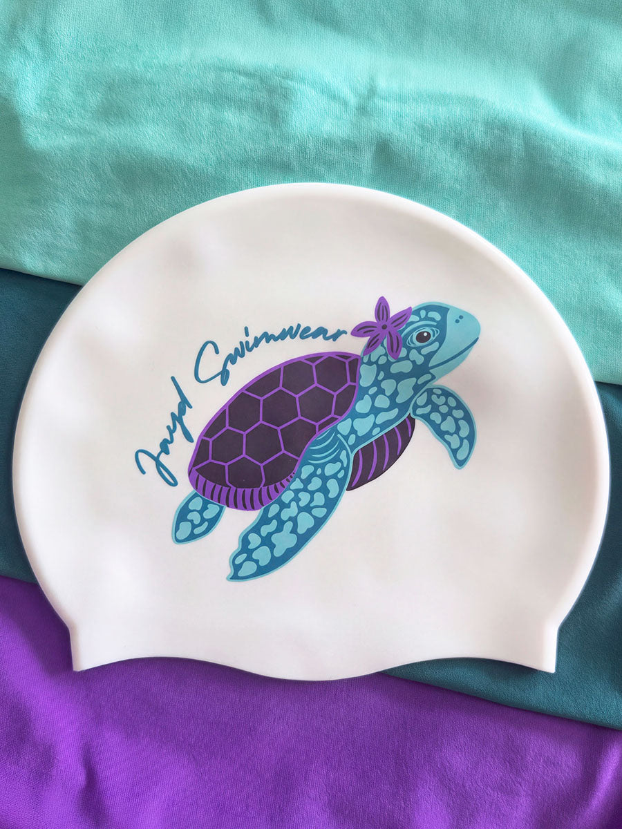A white silicone swim cap featuring a custom printed design of a purple and blue sea turtle with a purple flower on its shell, alongside the text 'Jazzy Swimswear'. The swim cap is laid flat on a background of teal and purple fabric.