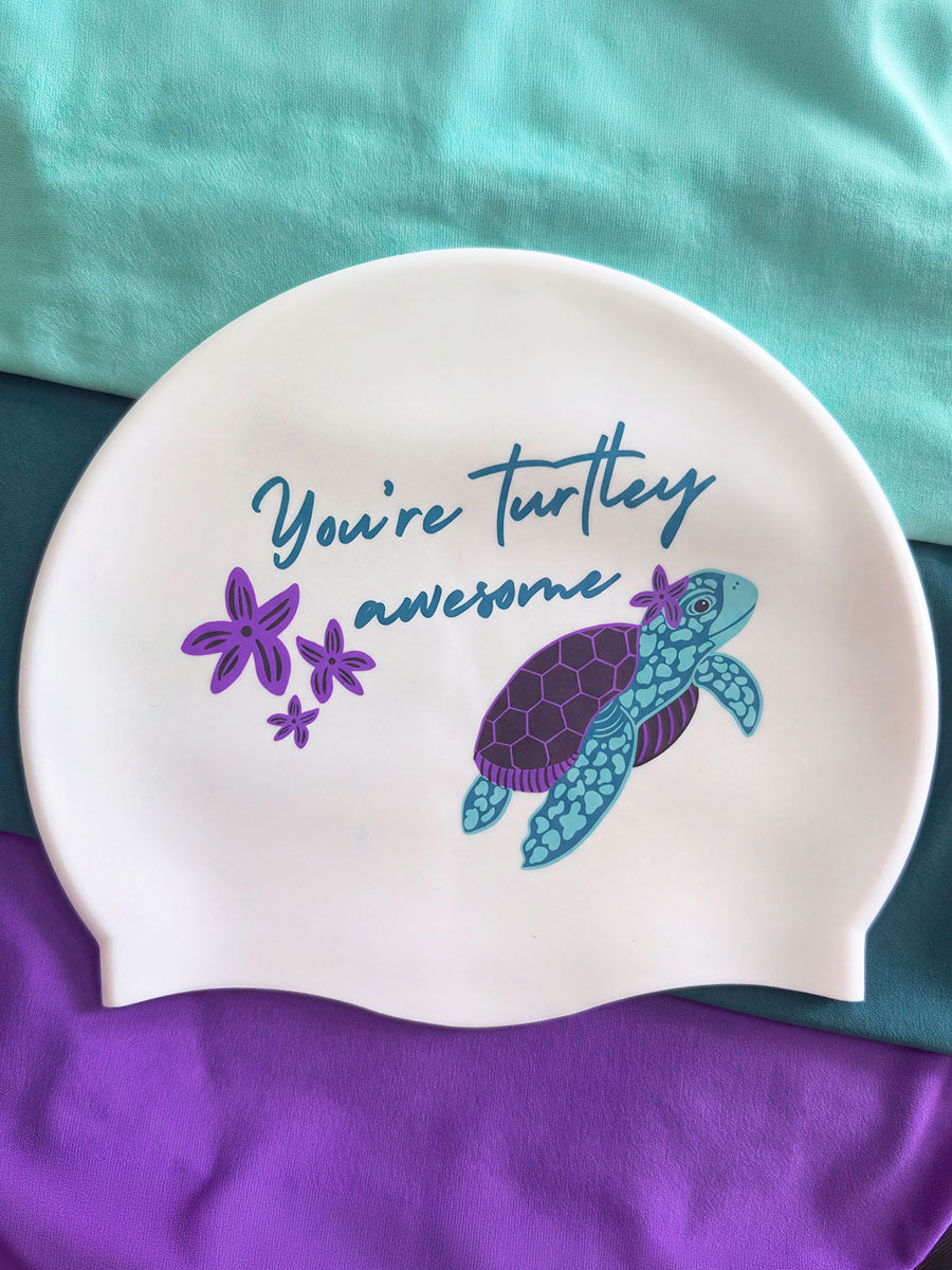 A white silicone swim cap featuring a blue and purple turtle illustration and the text 'You're turtle awesome' in blue script, displayed on a teal and purple fabric background.