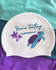 A white silicone swim cap featuring a blue and purple turtle illustration and the text 'You're turtle awesome' in blue script, displayed on a teal and purple fabric background.
